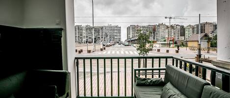 A balcony with a sofa and a wooden table, overlooking a street with buildings and a pedestrian crossing.