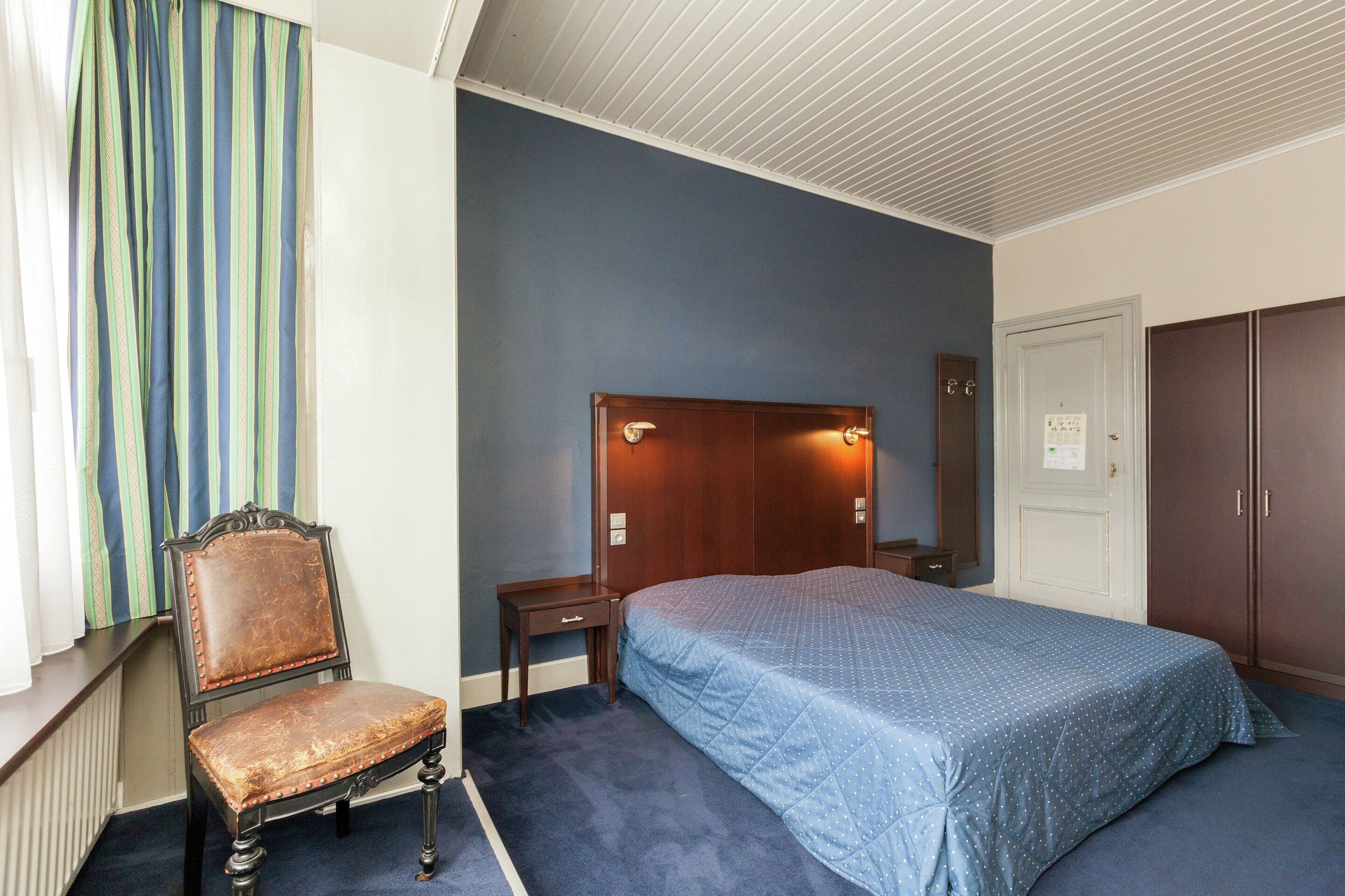 A bedroom with a bed, a chair, a nightstand, a wardrobe, and a window with curtains.