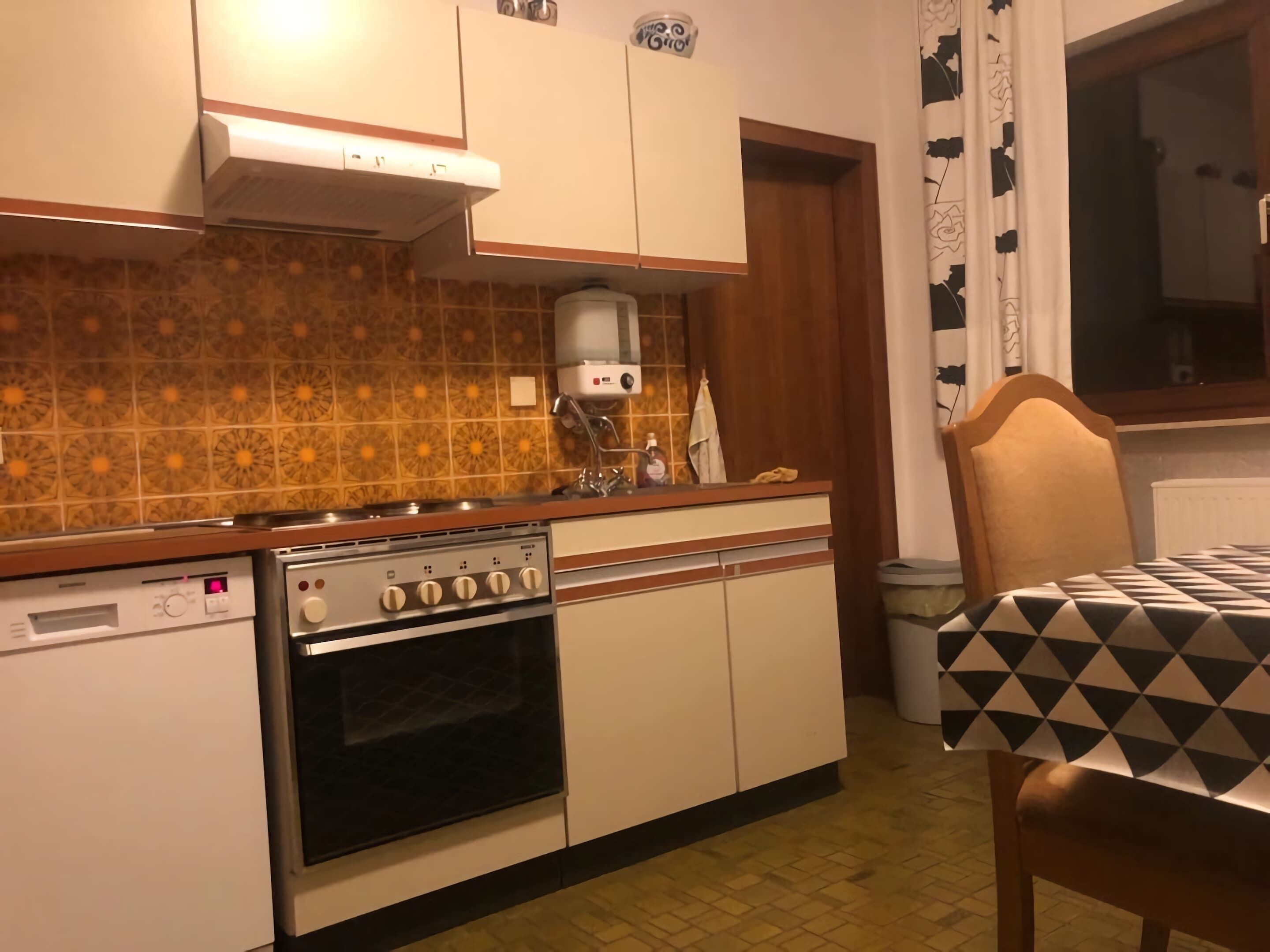 Een keuken met een fornuis, oven en wastafel, een stoel en een raam met gordijnen.