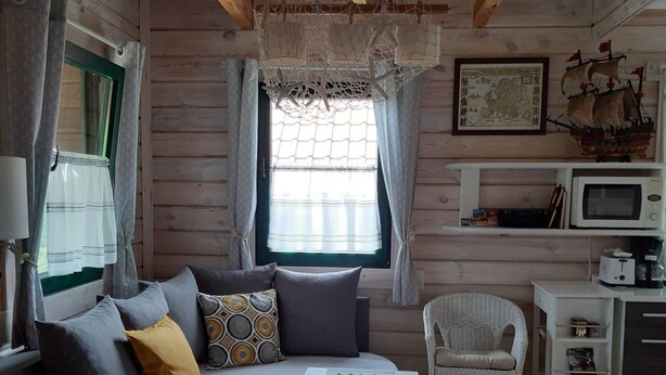 A cozy cabin-style room with a wooden interior, a sofa with cushions, a wicker chair, a small table, a window with curtains, a framed map, and a microwave on a shelf.