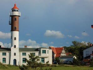 Un phare avec un sommet rouge, une base blanche et un petit balcon. Au premier plan, on aperçoit des bâtiments blancs avec des fenêtres vertes.