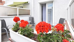 A balcony with a white planter containing red flowers, a grey chair, and a window with a curtain.