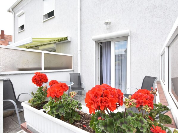 A balcony with a white planter containing red flowers, a grey chair, and a window with a curtain. - Tolles Ferienhaus in Wismar fur 8 Personen (Wismar)