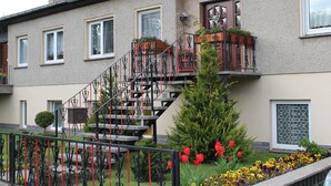A two-story building with a staircase, a small garden with flowers, and a metal fence.