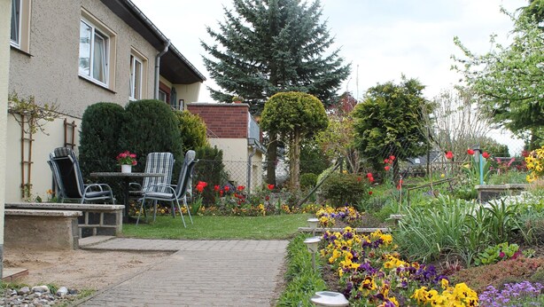 A residential garden with a paved walkway, a patio with chairs, and various flowering plants.