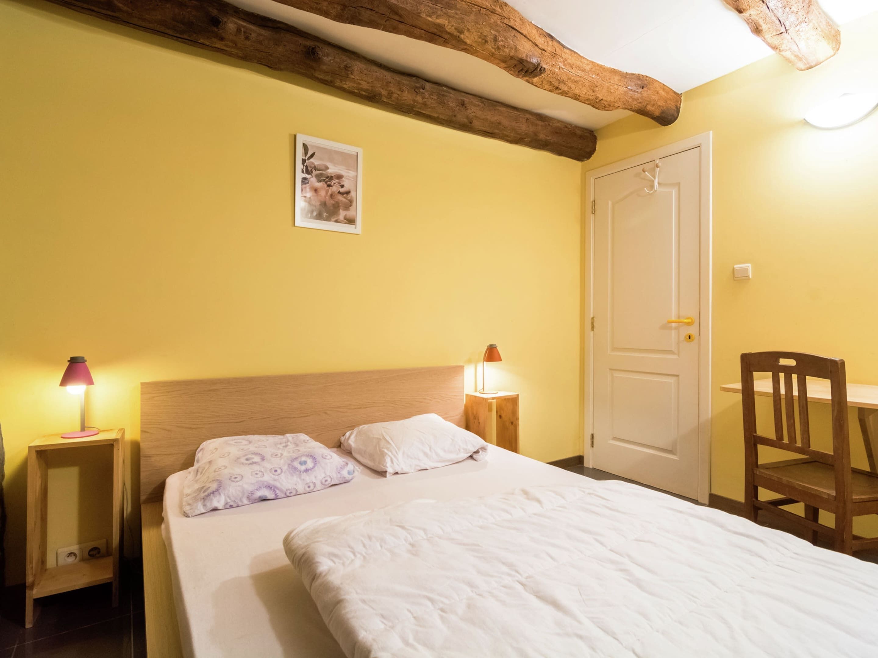 A bedroom with a wooden headboard, a bed with white bedding, two bedside lamps, a wooden chair, and a door.