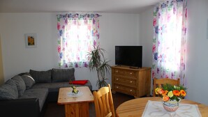 Un salon comprenant un canapé, un téléviseur, une table en bois avec des chaises, et une petite table sur laquelle est posé un vase de fleurs.
