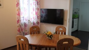 Un coin repas avec une table en bois et quatre chaises, un téléviseur à écran plat fixé au mur et un rideau à motifs floraux.