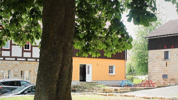 Une rue bordée d’arbres, avec un bâtiment en pierre, une voiture garée et une table de pique-nique.