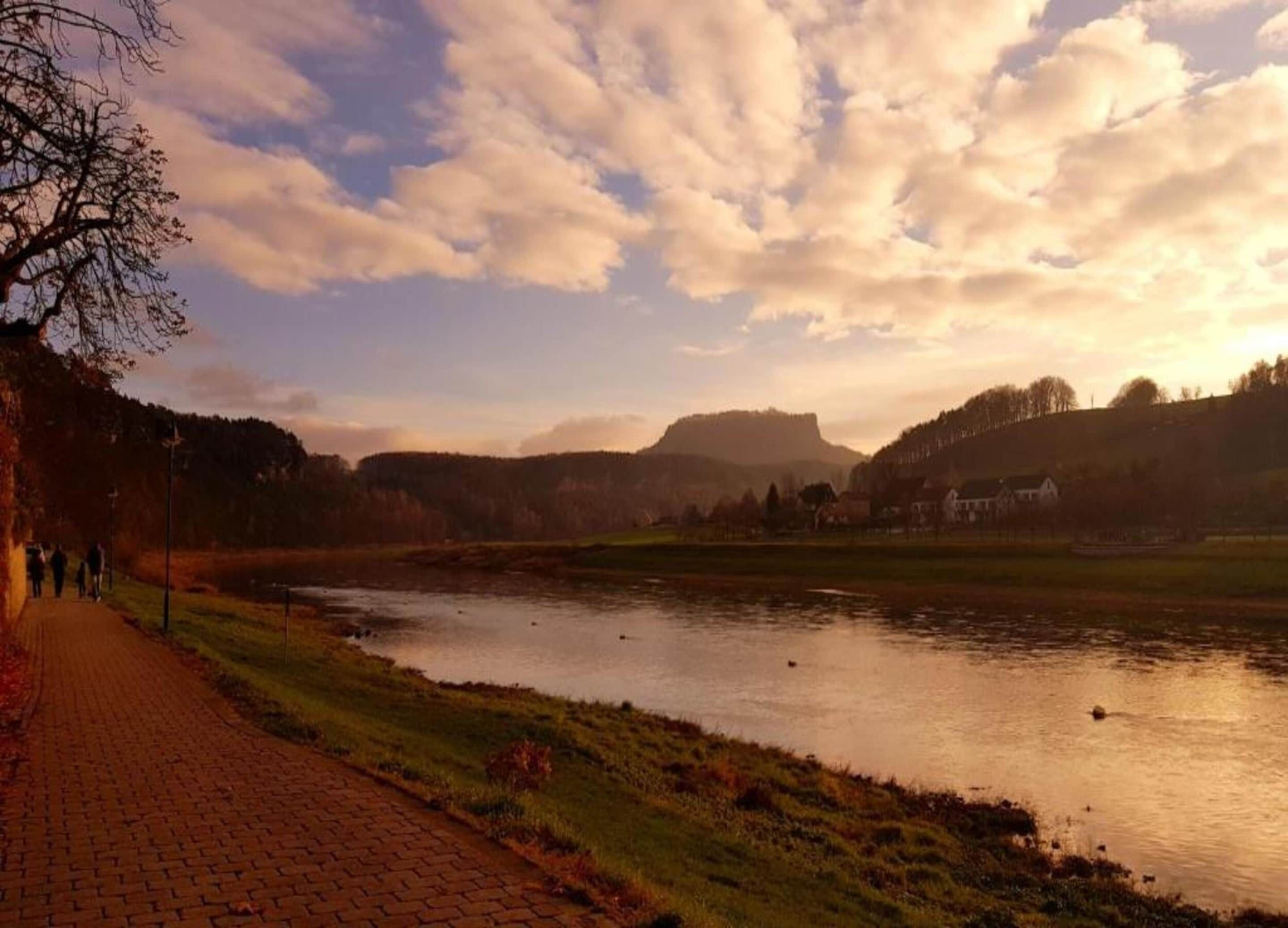A riverside path with a brick walkway, a calm river, and a backdrop of hills and a few buildings.