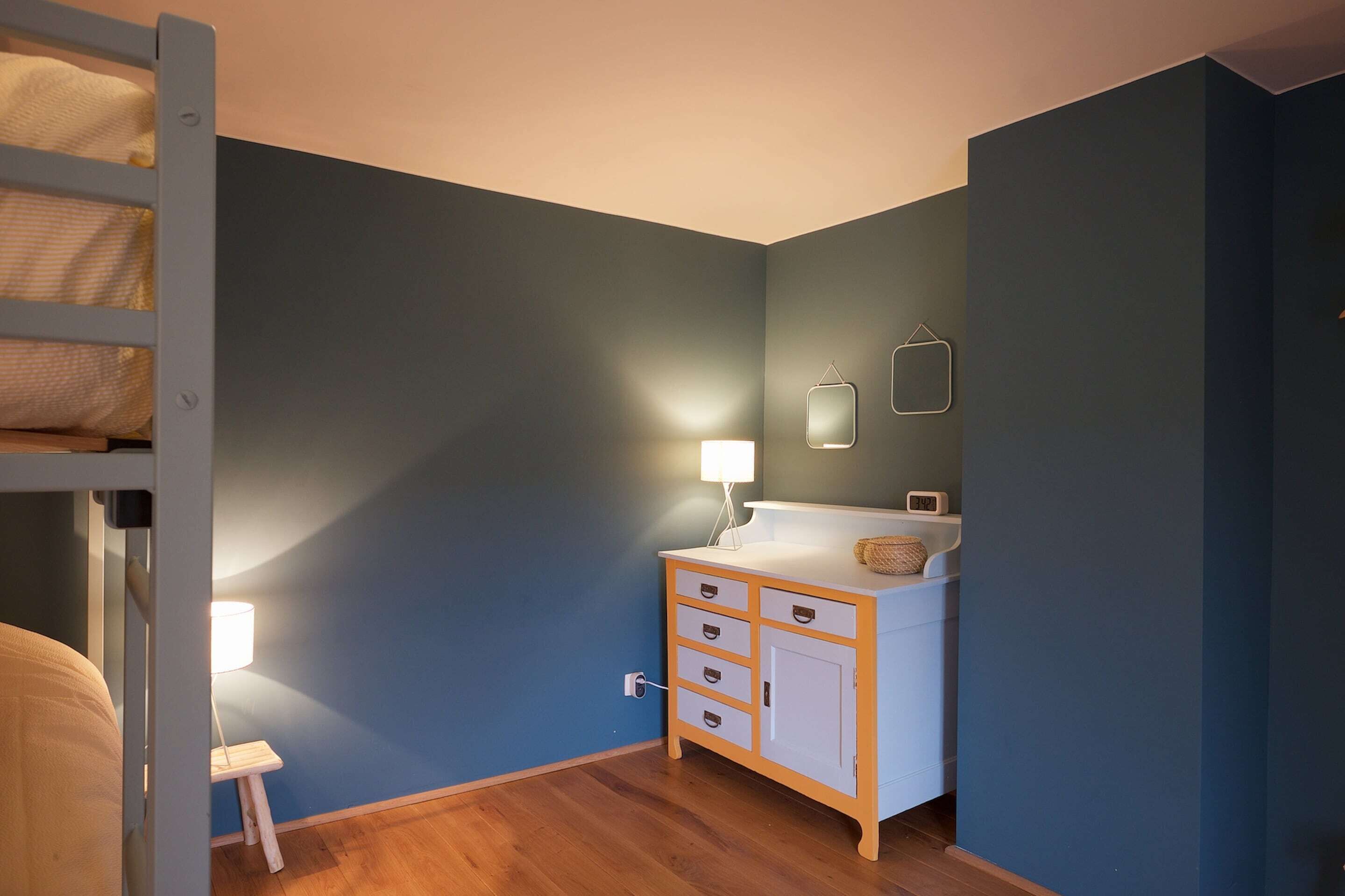 A bedroom with a bunk bed, a bedside table with a lamp, a dresser with drawers, and a wall-mounted mirror.