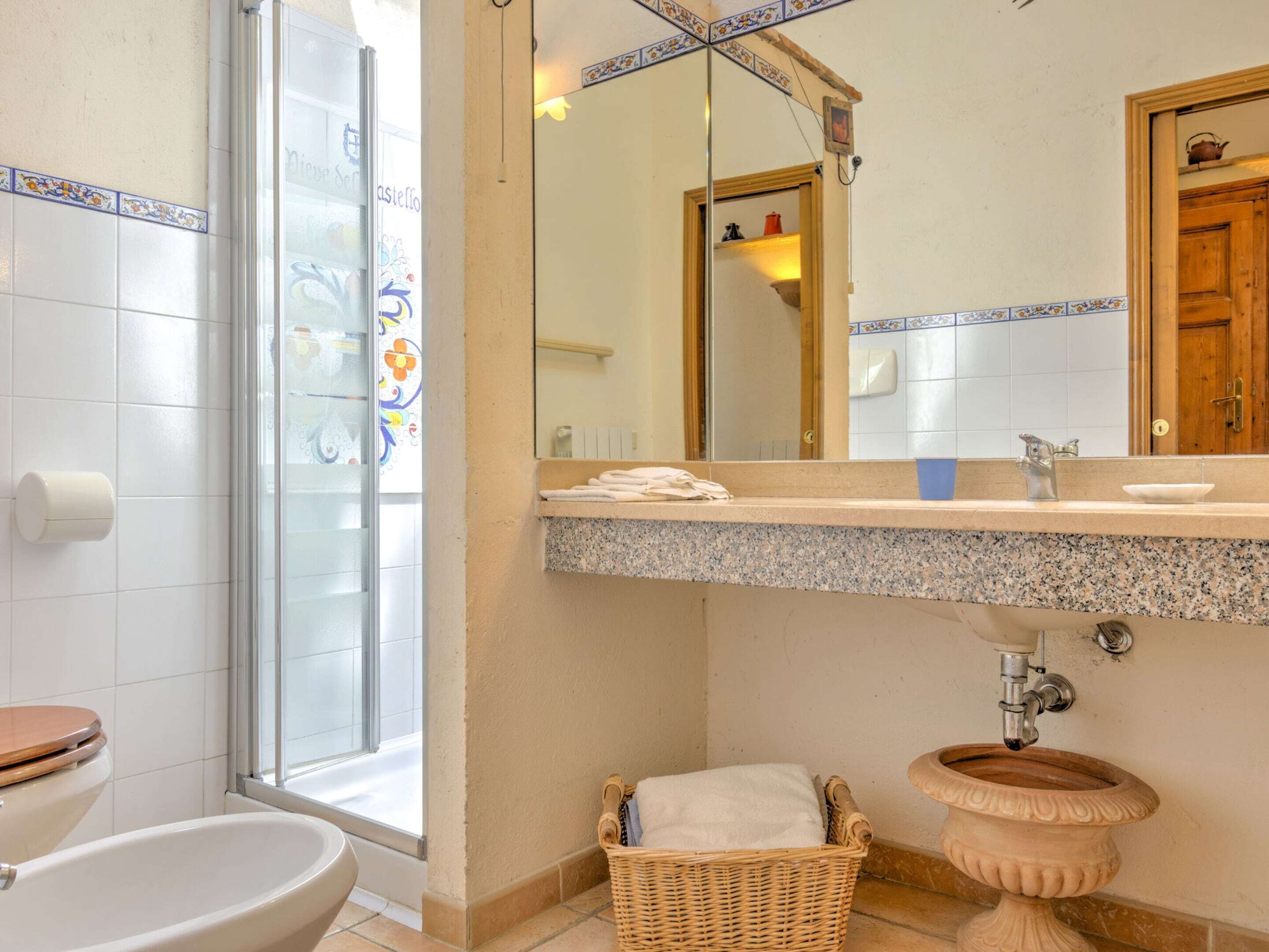 A bathroom with a toilet, a sink with a marble countertop, a decorative basin, a wicker basket, and a shower with frosted glass doors.