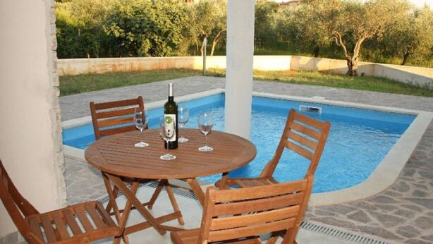 A round wooden table with four chairs around a pool area, with a bottle of wine and glasses on the table. - Holiday Home in Novigrad With Private Pool (Novigrad)