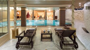 Un área de piscina interior lujosa con techo de cristal, tumbonas de madera y una zona de estar central con mesa y sillas.