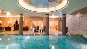 Un área de piscina interior lujosa, con suelo de baldosas, un mural en el techo y una zona de estar con un banco y una mesa.