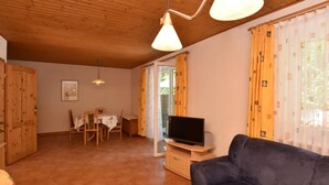 A cozy living room with a dark sofa, a wooden dining table with chairs, a television on a wooden stand, and a large window with curtains. - Ferienhaus in Damshagen Nahe Ostsee (Damshagen)