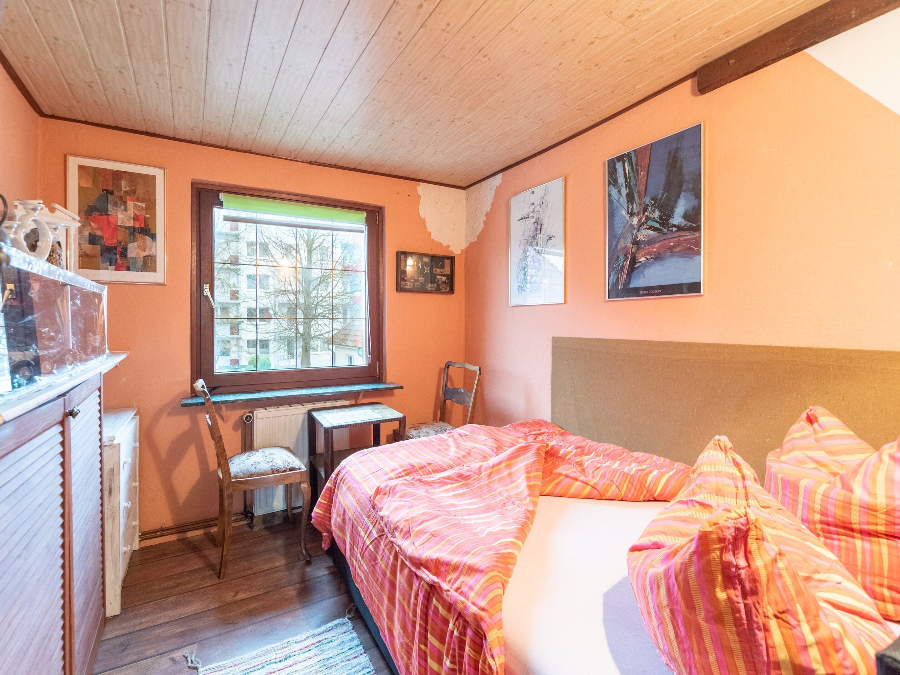 A bedroom with a wooden ceiling, a bed with striped bedding, a desk with a chair, and a window with a view of buildings.