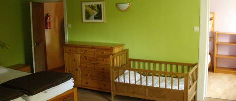 A room with a wooden crib, a wooden dresser, and a bed with a dark cover.