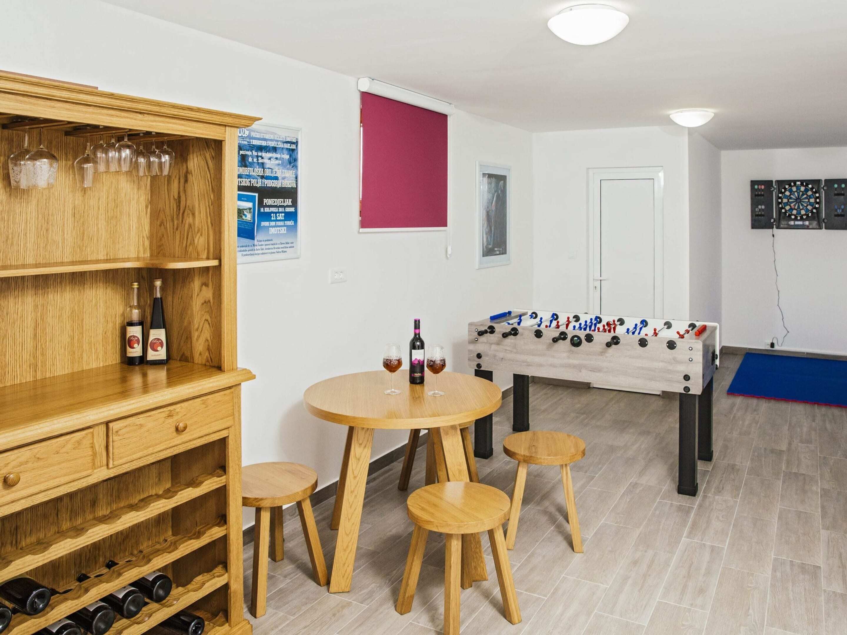 A room with a wooden table, four stools, a foosball table, and a wine rack with bottles.