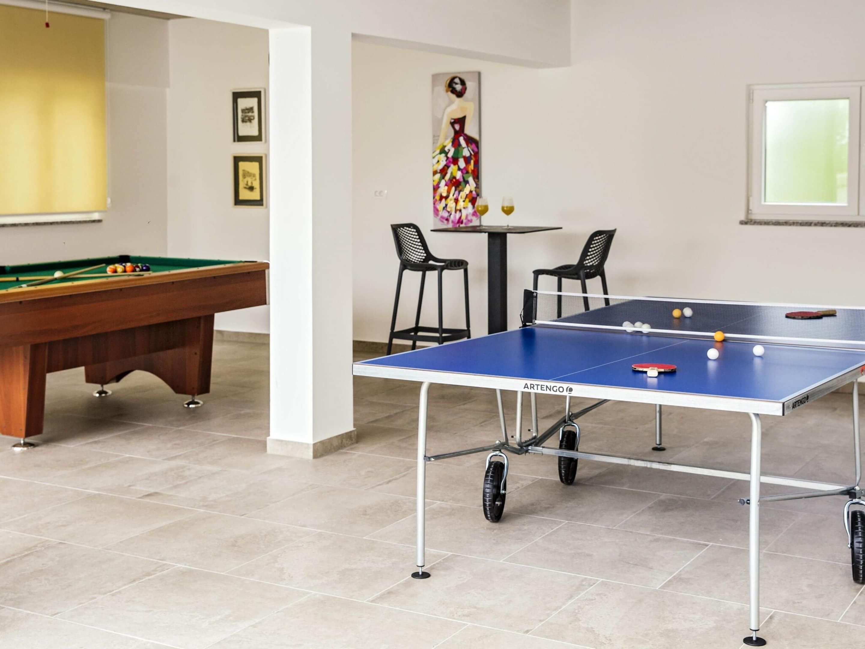 A pool table, a ping pong table, and two chairs in a room with tiled flooring and a painting on the wall.