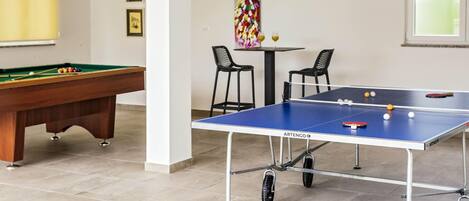 A pool table, a ping pong table, and two chairs in a room with tiled flooring and a painting on the wall.