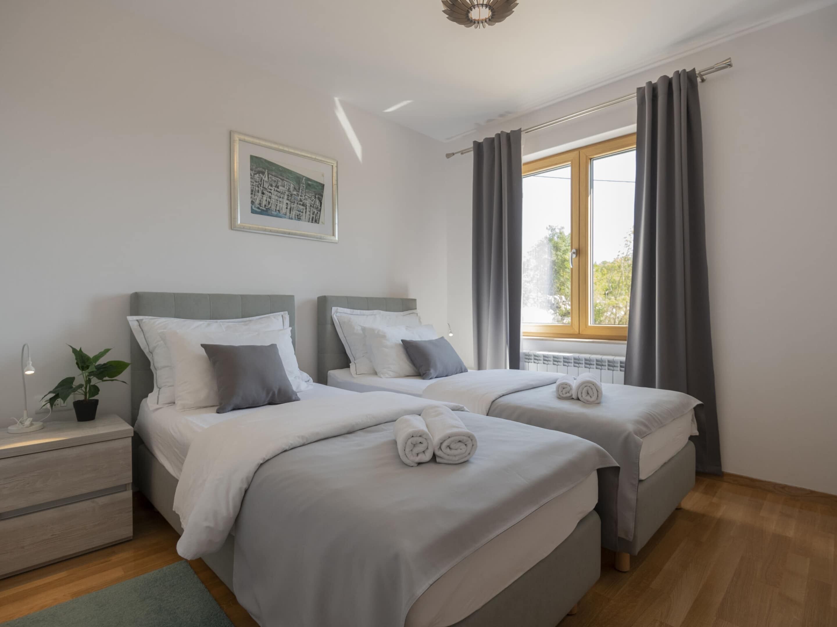 A modern bedroom with two beds, a nightstand with a lamp, a potted plant, and a window with curtains.