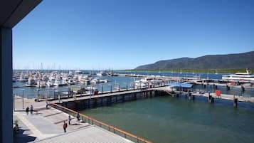A marina with multiple boats docked, a pier with people walking, and a hilly landscape in the background.