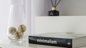Un florero de vidrio transparente con unas cuantas madejas de lana sobre una superficie blanca, junto a un libro titulado 'minimalism'.