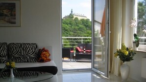 Un salón moderno con un sofá estampado con motivos de cebra, un ventanal de vidrio que da a un balcón con vistas a una colina y un edificio, y un jarrón con flores amarillas sobre una mesa.