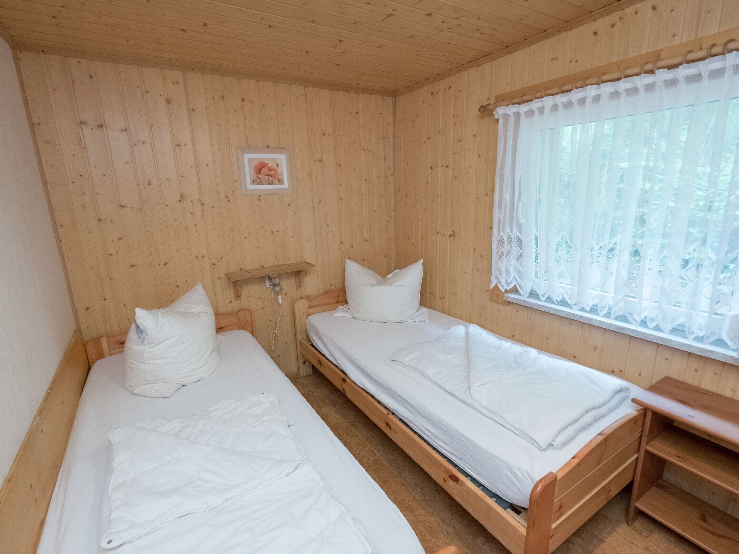 A wooden cabin room with two single beds, a small shelf, and a window with lace curtains.