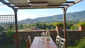 Een houten tafel met stoelen onder een pergola, met uitzicht op een schilderachtig landschap met heuvels en een verafgelegen stad.