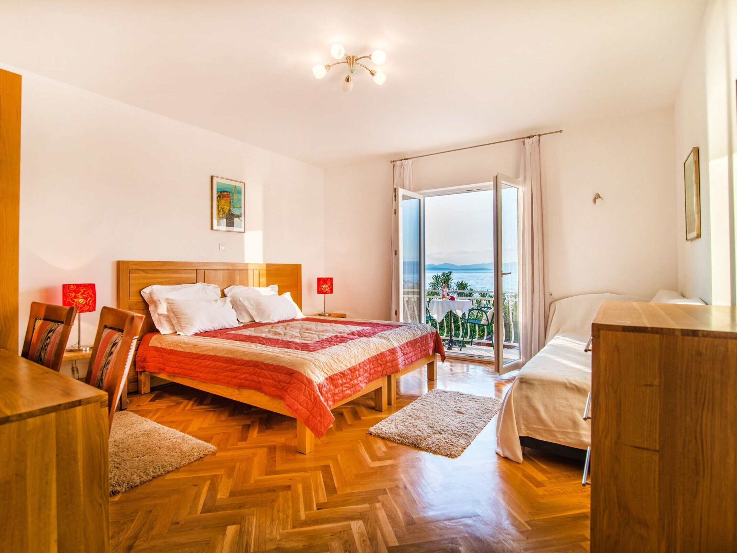 A hotel room with a wooden floor, a bed with a red and white quilt, a desk with a chair, a small table with a chair, and a balcony with a view of the sea.
