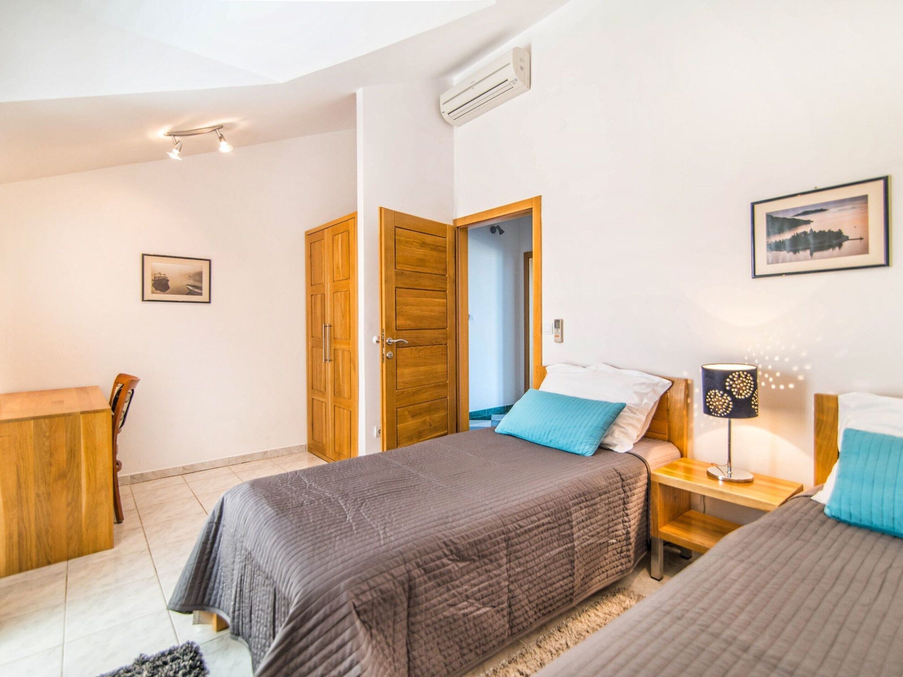 Un dormitorio con dos camas, un armario de madera, un escritorio, una silla, una mesita de noche con lámpara, y un cuadro en la pared.