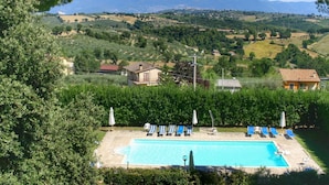 A swimming pool with lounge chairs and umbrellas, surrounded by greenery and houses, with mountains in the background. - Farmhouse Bliss With Pool, Wine and Tails (Gualdo Cattaneo)
