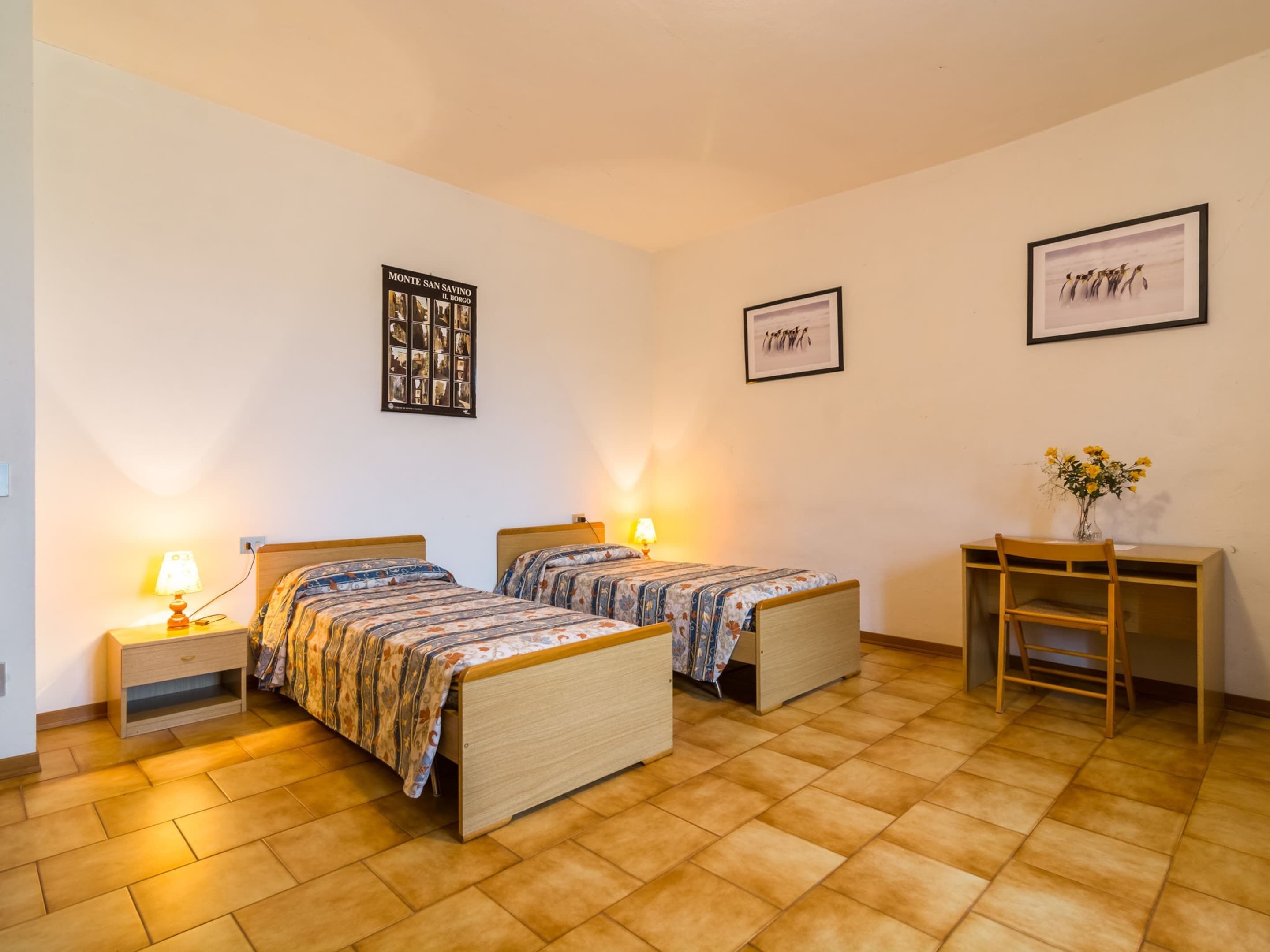 A room with two beds, a desk, and a chair. There are two lamps on the nightstands.