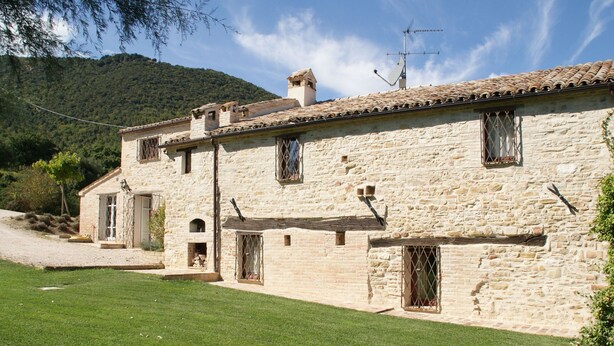 A stone building with multiple windows and a door, surrounded by a well-maintained lawn and a mountain in the background. - Belvilla by OYO Appartamento dei Cavalieri (Treia)
