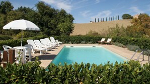 A swimming pool with lounge chairs and an umbrella, surrounded by greenery and a hillside. - Belvilla by OYO Appartamento dei Cavalieri (Treia)