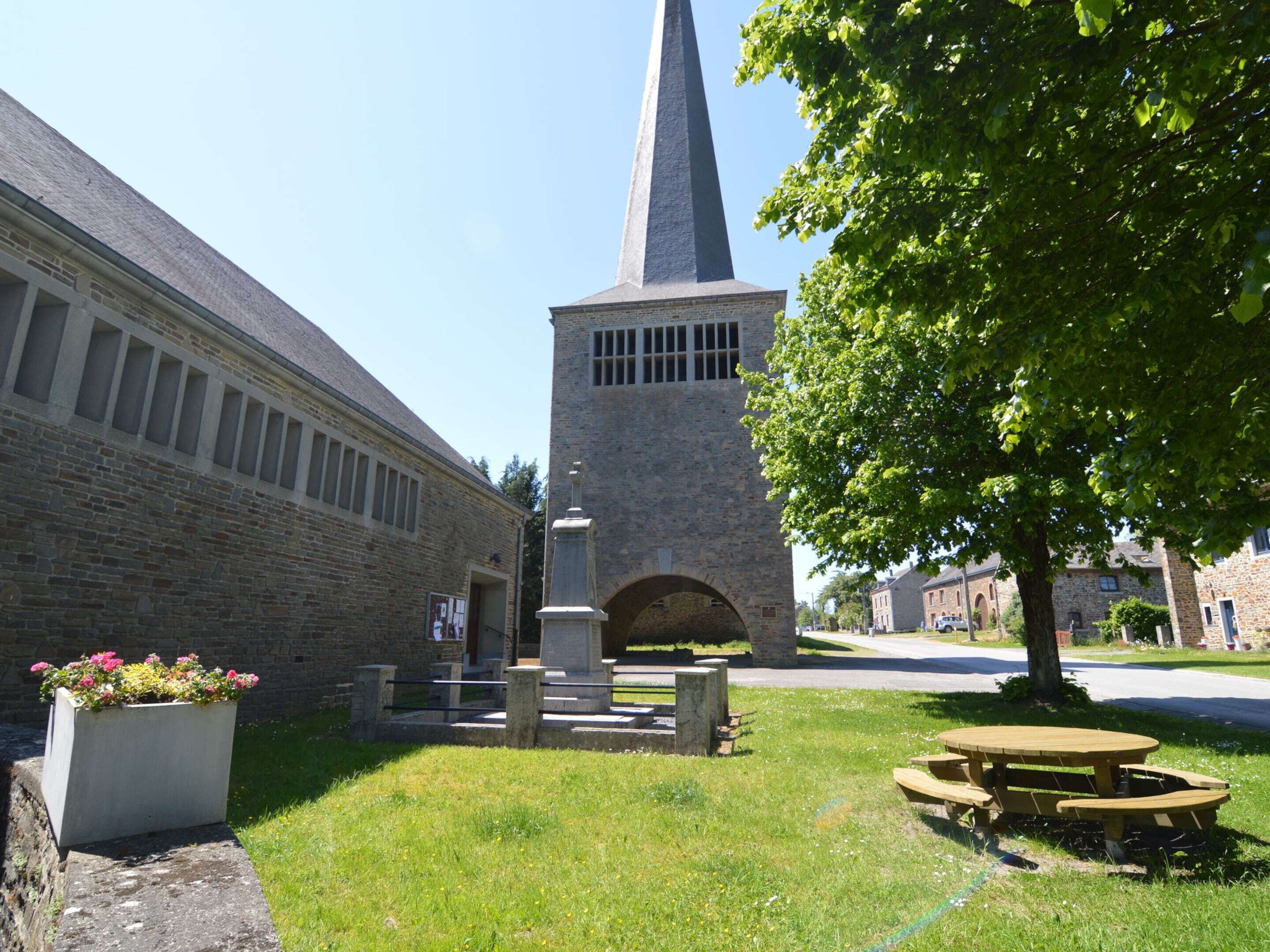Un cortile con un edificio in pietra, caratterizzato da una torre alta, un aiuola fiorita e un tavolo da picnic.