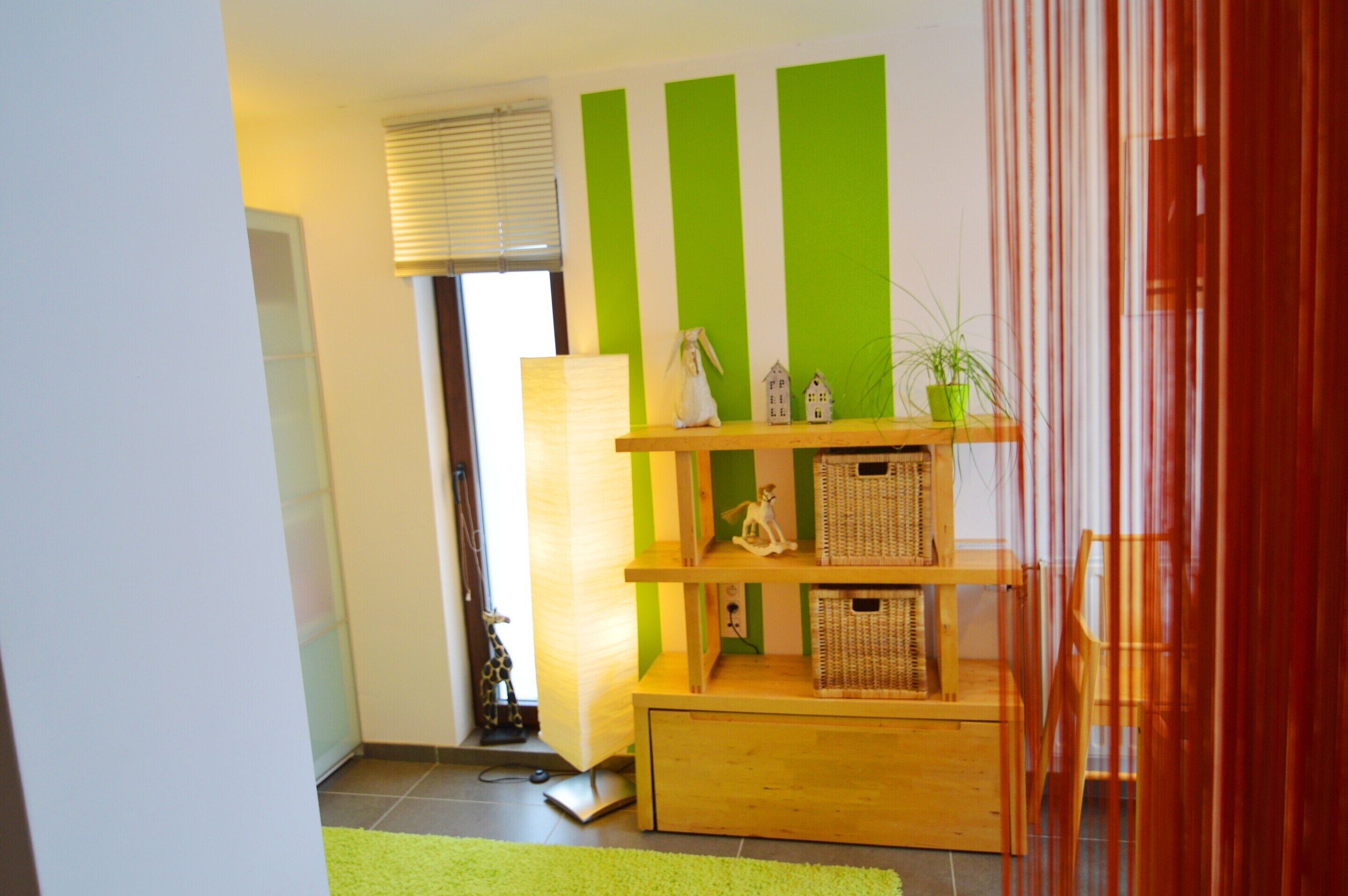 A room with a wooden shelf unit displaying decorative items, a green-striped wall, and a window with blinds.