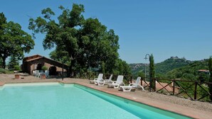 Ein Swimmingpool mit Liegestühlen und einem kleinen Gebäude, umgeben von Bäumen und mit Blick auf einen Hügel mit einer schlossartigen Struktur.