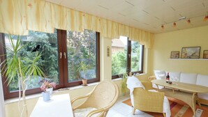 A room with wicker furniture, a white table, and a sofa. There are windows with curtains, a potted plant, and a bottle of wine on the table. - Apartment in Harz With Terrace (Ballenstedt)