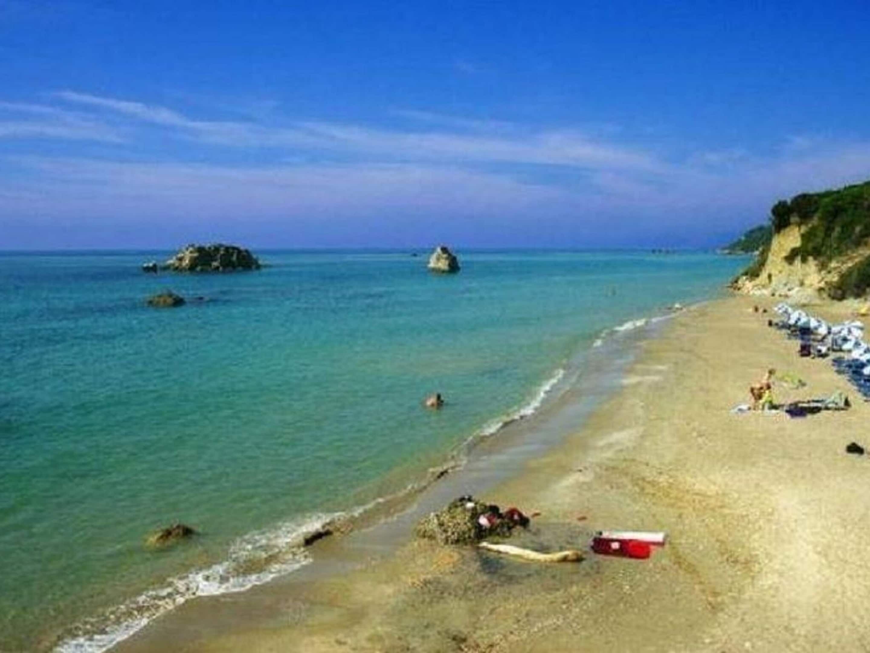 Une plage avec une eau turquoise et limpide, des affleurements rocheux et une rive sablonneuse parsemée de parasols et de serviettes de plage.