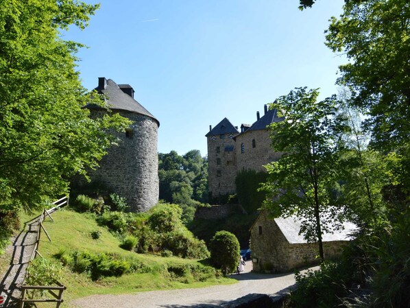 Um castelo de pedra histórico com uma torre redonda e um edifício maior com telhados inclinados, cercado por vegetação exuberante e um caminho de cascalho.