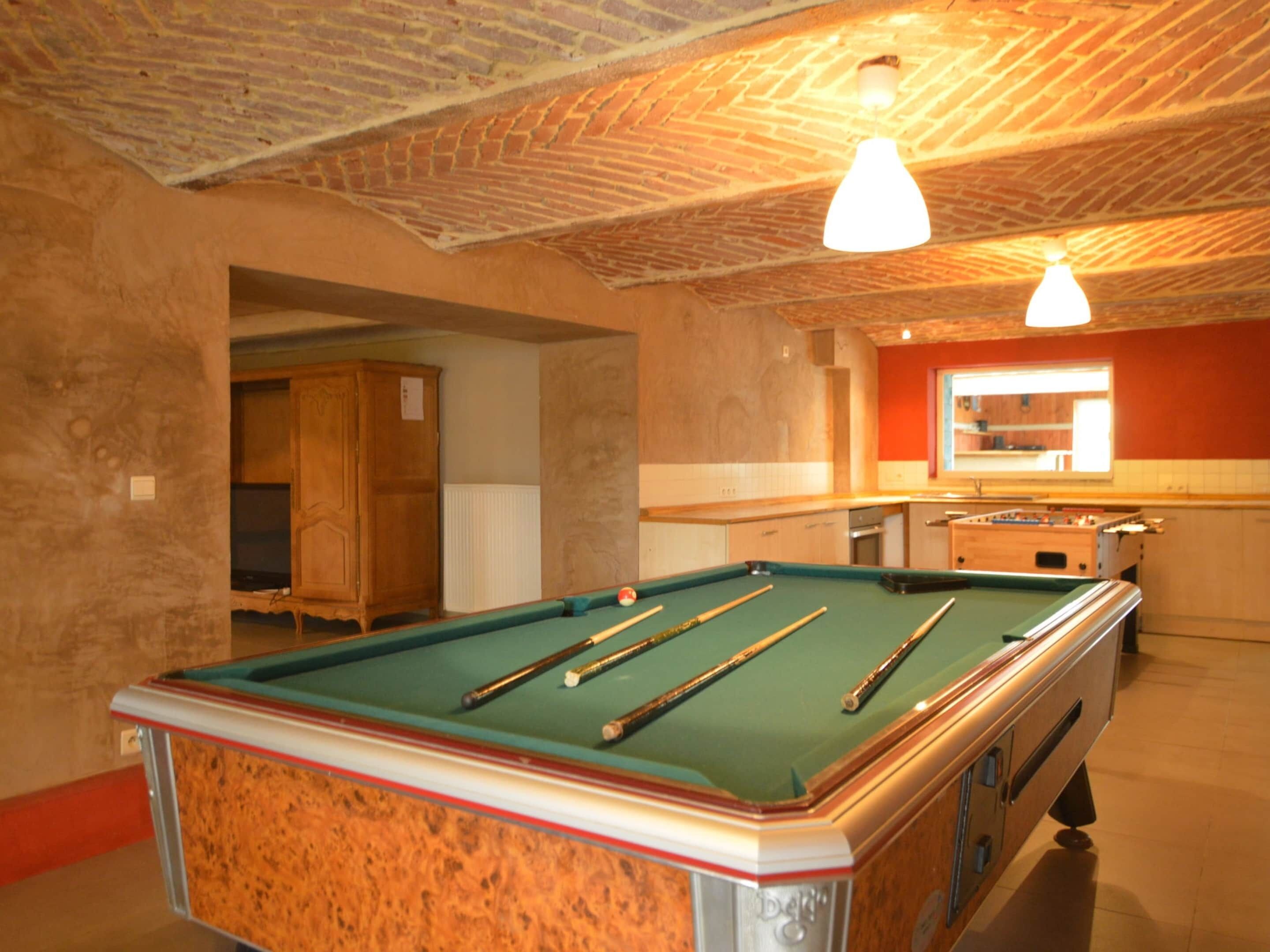 A pool table with cues, a foosball table, and a kitchen area with a window.