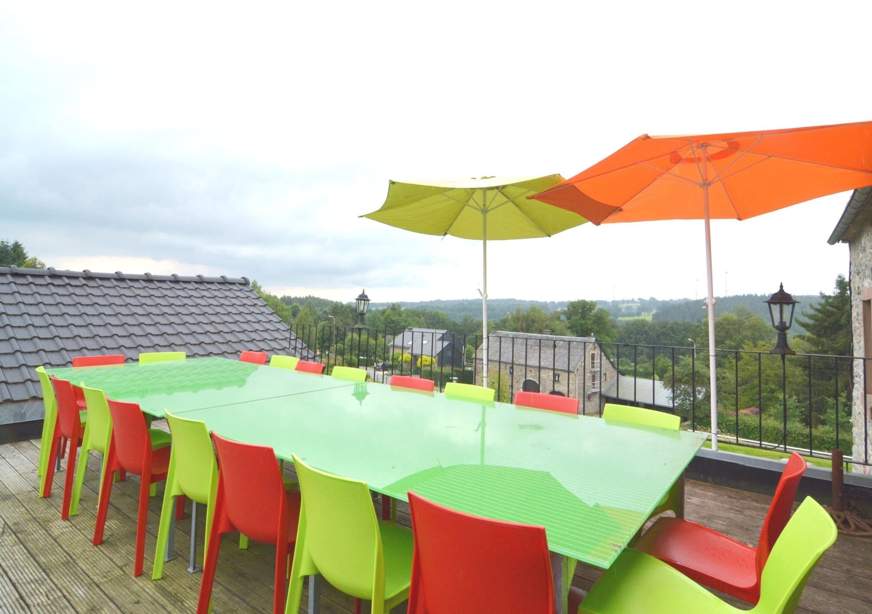 Una mesa verde con sillas rojas en una terraza exterior con vistas a edificios y árboles.