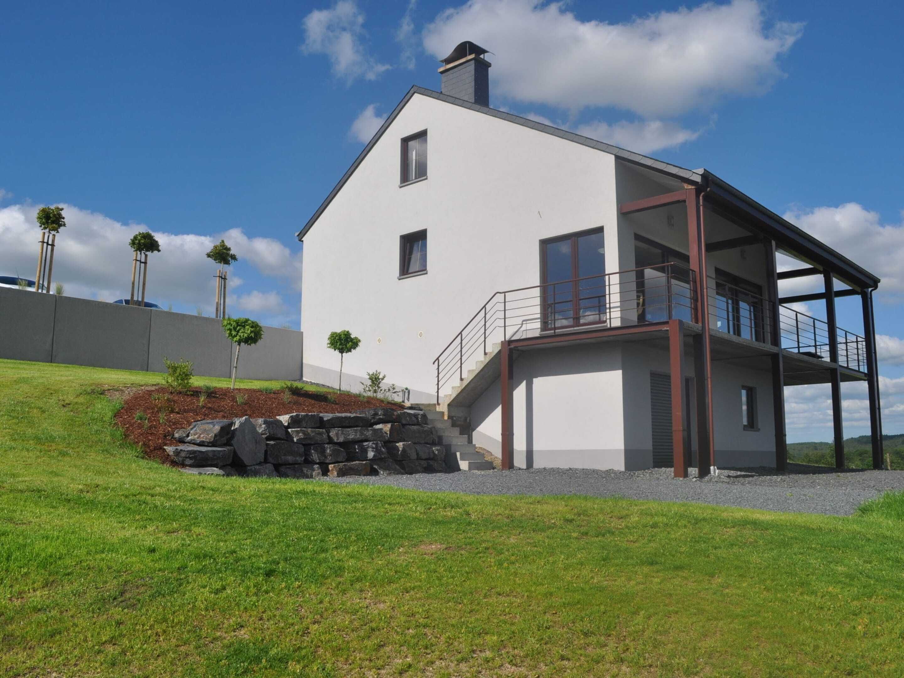 A modern two-story house with a balcony, a stone retaining wall, and a grassy lawn.