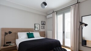 Une chambre d’hôtel moderne avec une tête de lit en bois, un grand lit avec des oreillers blancs et verts, un pouf noir, une lampe sur pied et une grande fenêtre avec des rideaux.