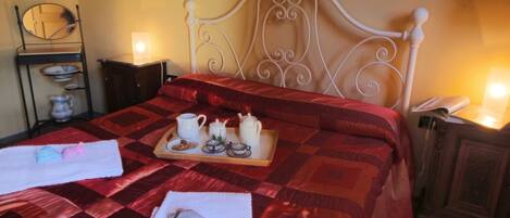 A neatly made bed with a red quilt, a tray with tea and pastries, and a bedside table with a lamp and a phone.