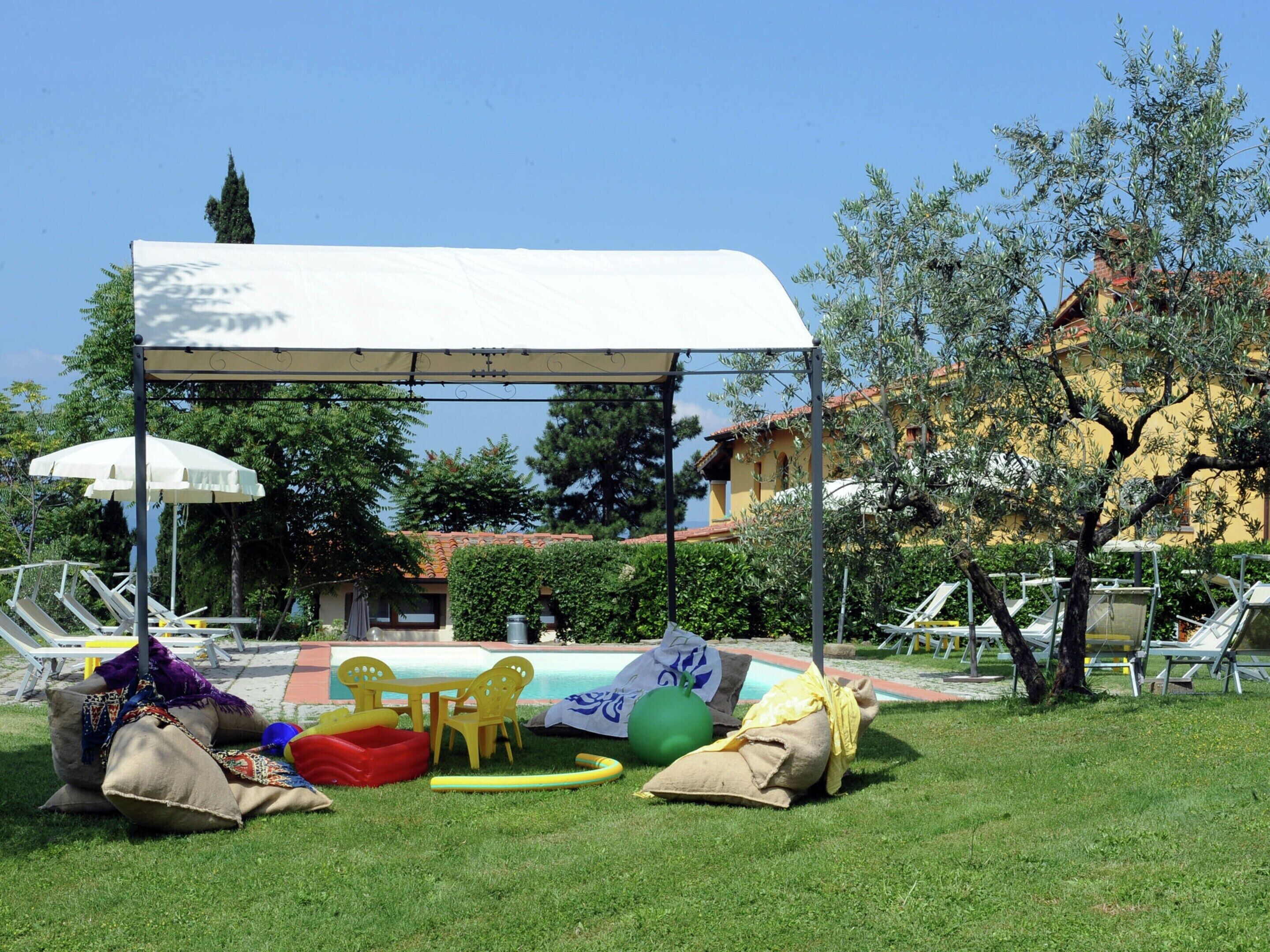 A covered outdoor seating area with chairs and tables, a pool in the background, and a building with a tiled roof.