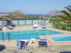 Ein Poolbereich mit Liegestühlen und Sonnenschirmen, eine Steinmauer und Blick auf das Meer und die Berge.